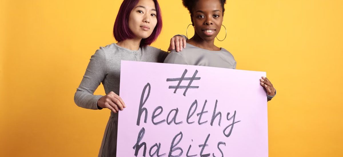 Two women holding a sign with '#healthy habits' on a vibrant yellow background.